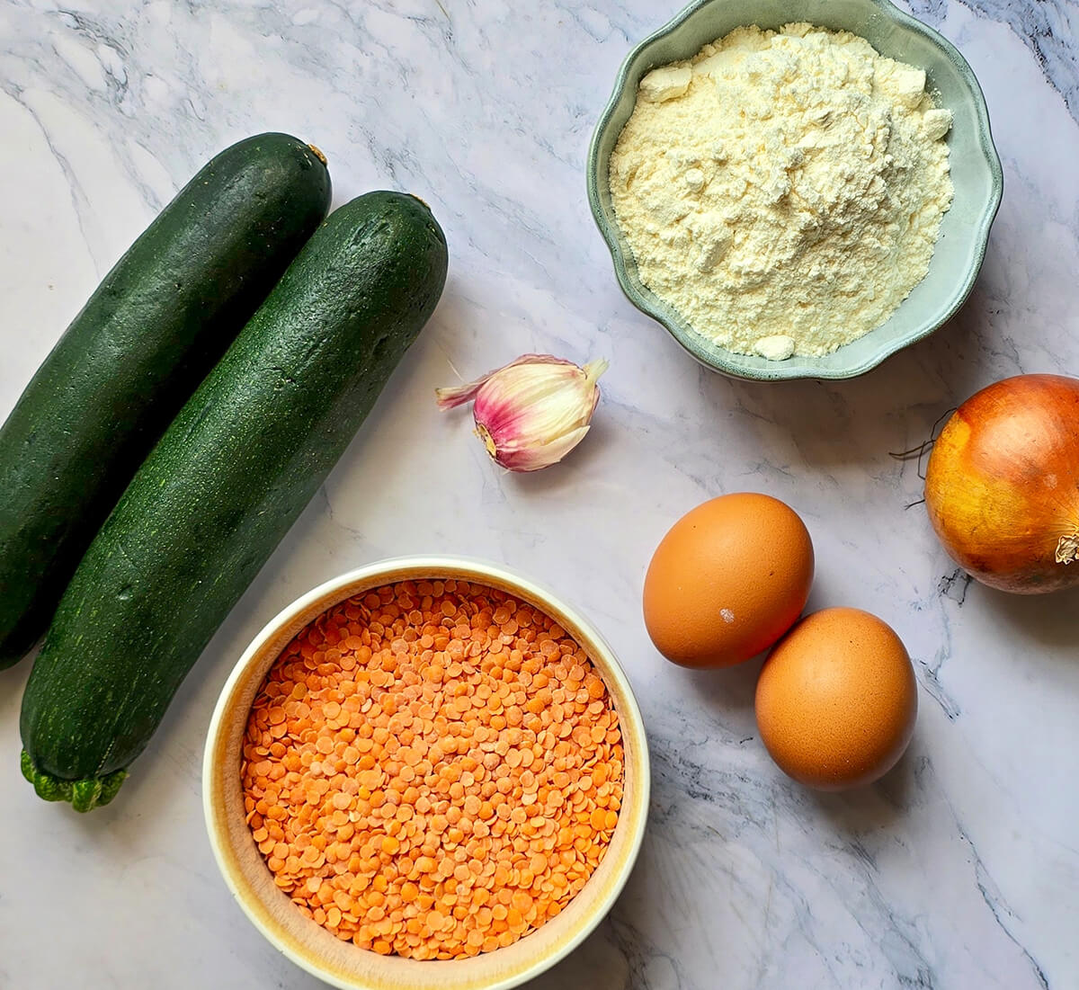 Ingrédients pour galettes de courgettes et lentilles corail disposés sur un plan de travail en marbre : deux courgettes vertes, un bol de lentilles corail, de la farine, deux œufs, un oignon et une gousse d'ail.