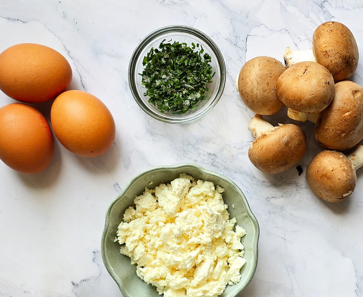 Des ingrédients pour faire une omelette champignons feta, sur un fond blanc marbré.