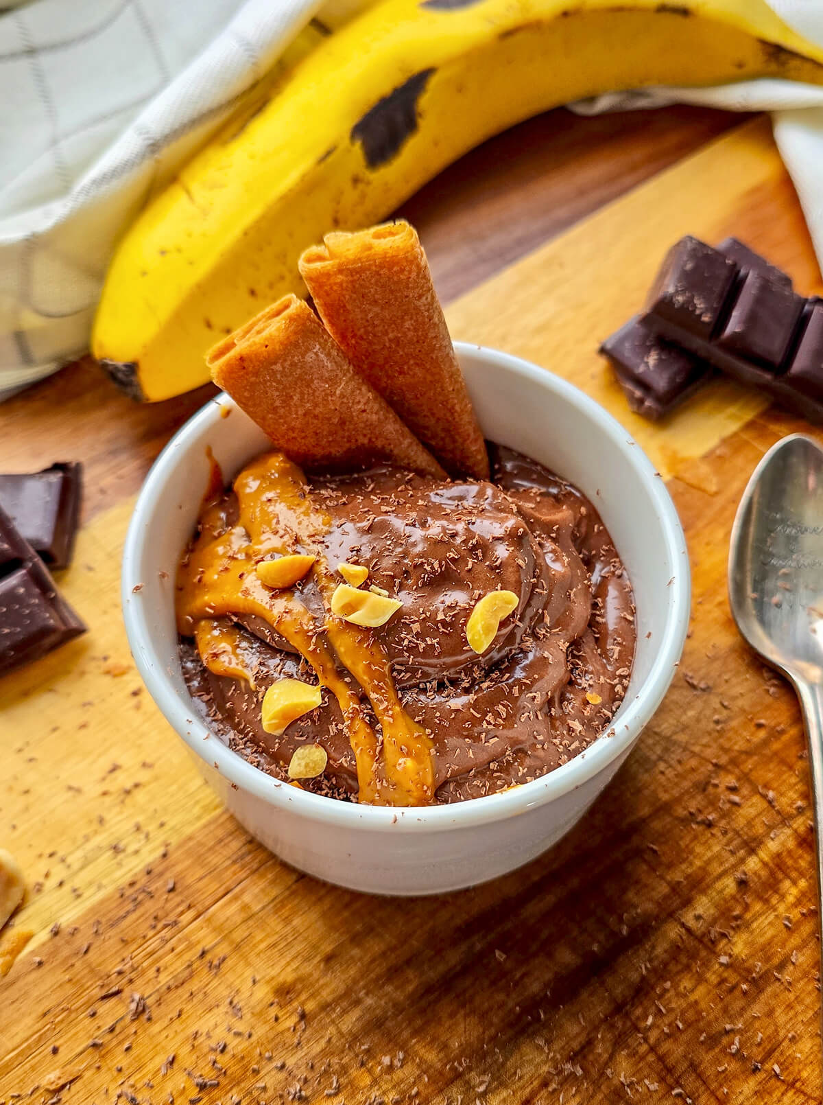 Recette healthy de glace banane chocolat maison sans sorbetière. Bol de nice cream onctueuse nappée de beurre de cacahuètes, parsemée d'éclats de cacahuètes et de copeaux de chocolat, présentée sur une planche en bois.