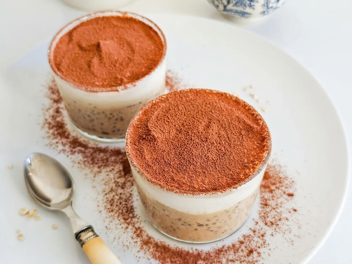 Vue de dessus de deux verrines d'overnight oats façon tiramisu, saupoudrées de cacao sur une assiette blanche avec une cuillère.