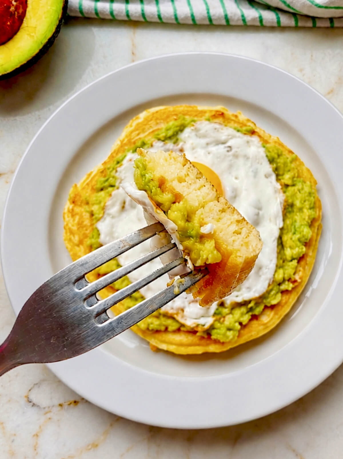 Une assiette avec un pancake salé XXL, garni d'avocat écrasé, et d'un oeuf sur le plat, une fourchette tenant un morceau de pancake.