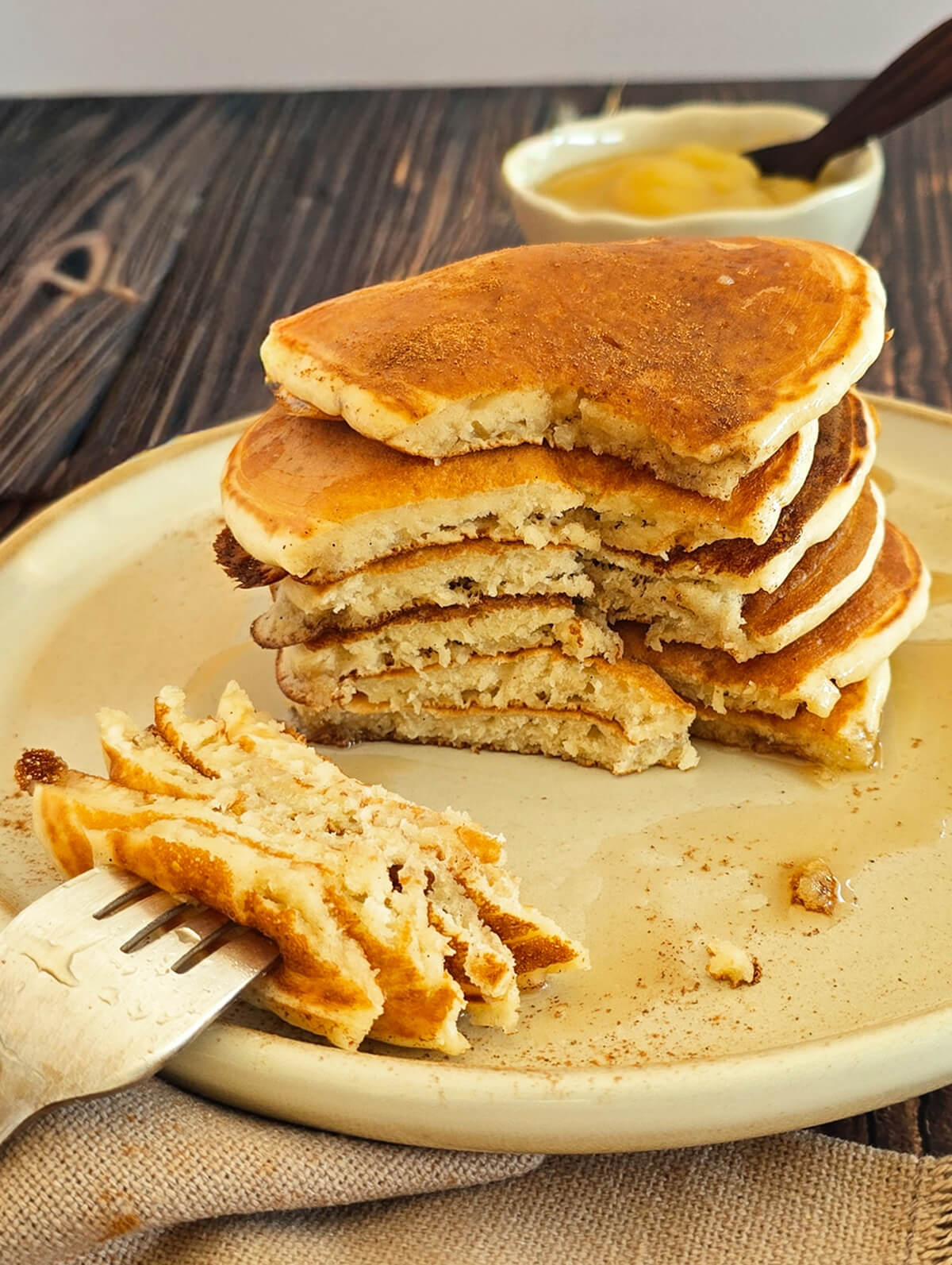 Une pile de pancakes sans œufs moelleux et dorés, coupés en deux pour montrer leur texture aérienne. Un morceau est piqué au bout d'une fourchette au premier plan, le tout nappé de sirop et saupoudré de cannelle sur une assiette claire.
