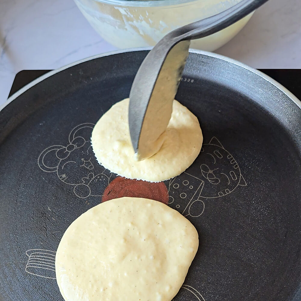 Une main versant une louche de pâte à pancakes dans une poêle noire anti-adhésive.