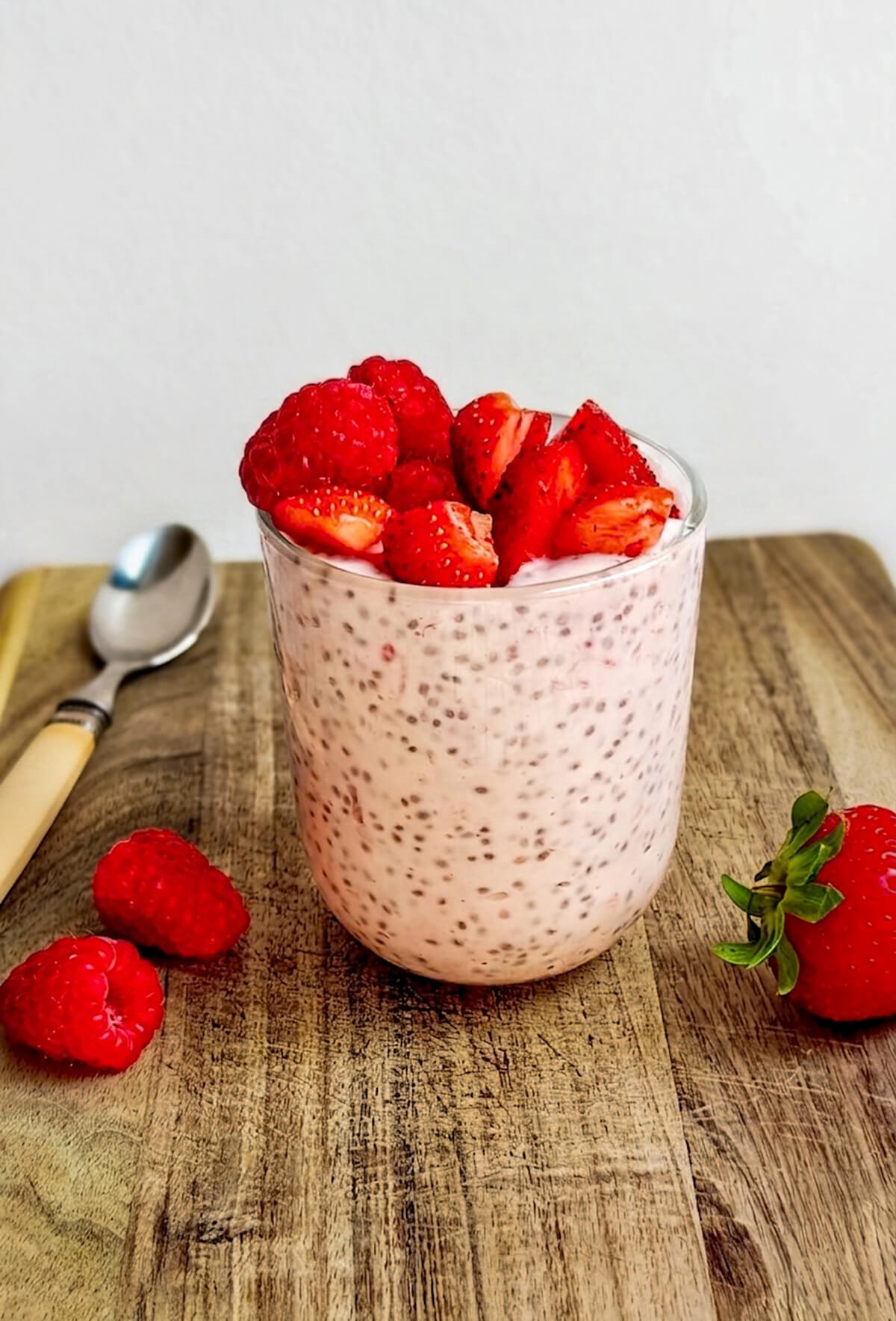 Pudding de chia à la fraise et framboise servi dans un verre transparent sur une planche à découper. Le verre est généreusement garni de fruits rouges frais. Une cuillère et quelques fruits entiers sont disposés en arrière-plan sur le bois rustique.