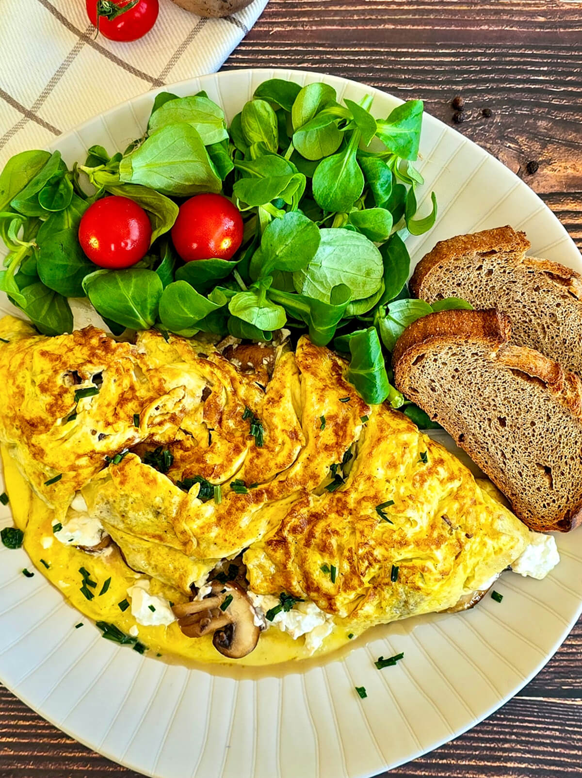 Une omelette aux champignons feta dans une assiette, avec des tranches de pain de campagne et une salade de mâche, sur un fond de bois.