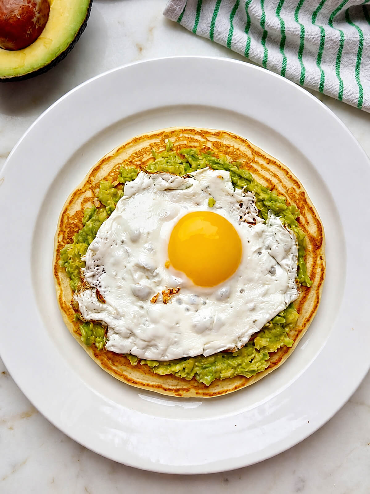 Un pancake salé dans une assiette blanche, garni d'avocat écrasé et d'un oeuf sur le plat, sur un fond blanc marbré, avocat et torchon en arrière plan.