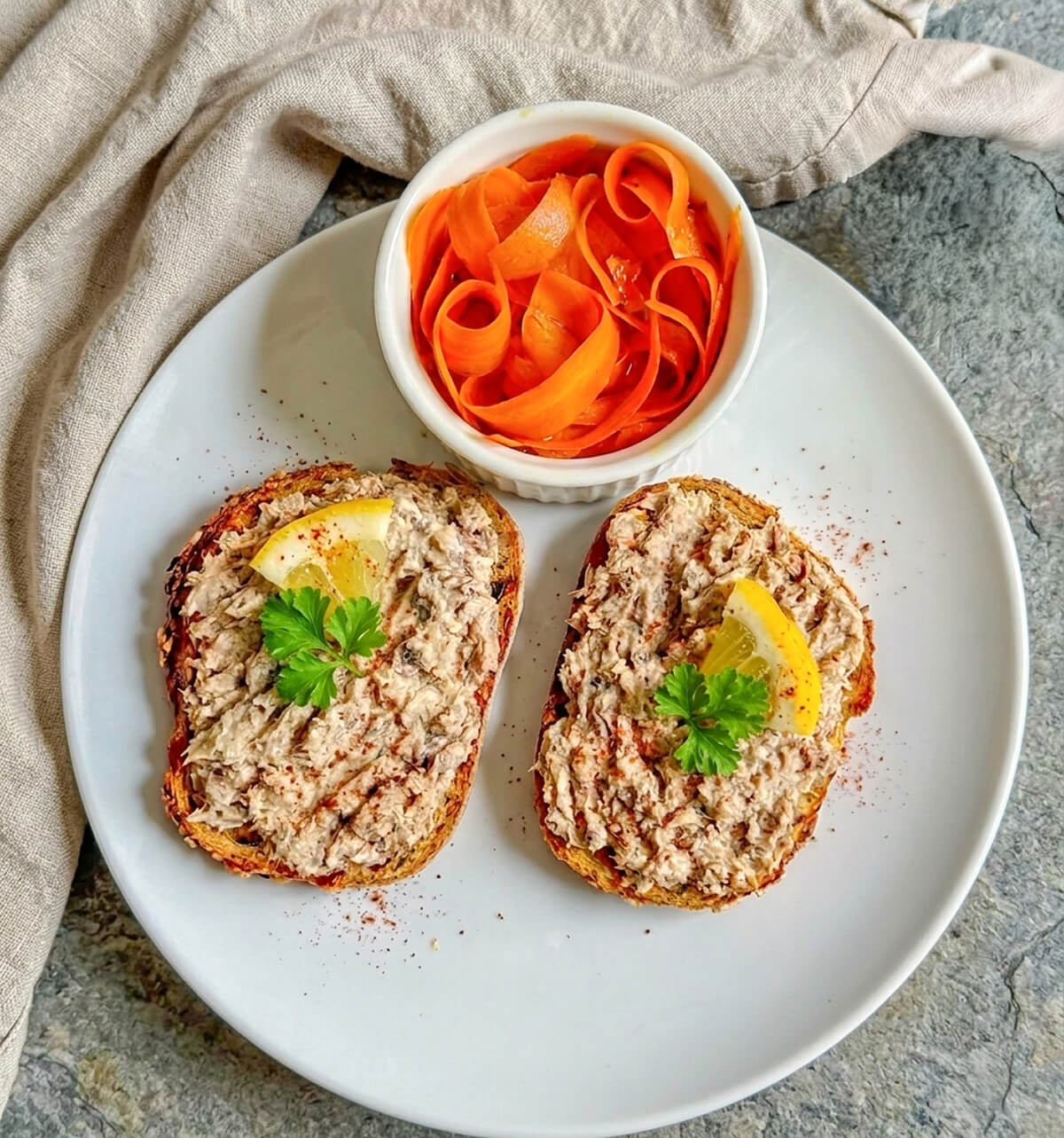 Vue du dessus d'une assiette blanche composée de deux tartines de rillettes de sardines sur du pain grillé, décorées d'une tranche de citron et de persil, accompagnées d'un ramequin de rubans de carottes.