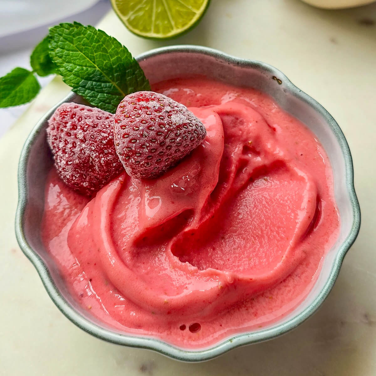 Recette de sorbet fraise maison facile et rapide à faire sans sorbetière. Vue de dessus d'un bol de glace à la fraise onctueuse, décoré de fraises givrées et de menthe fraîche, avec des citrons verts en arrière-plan.
