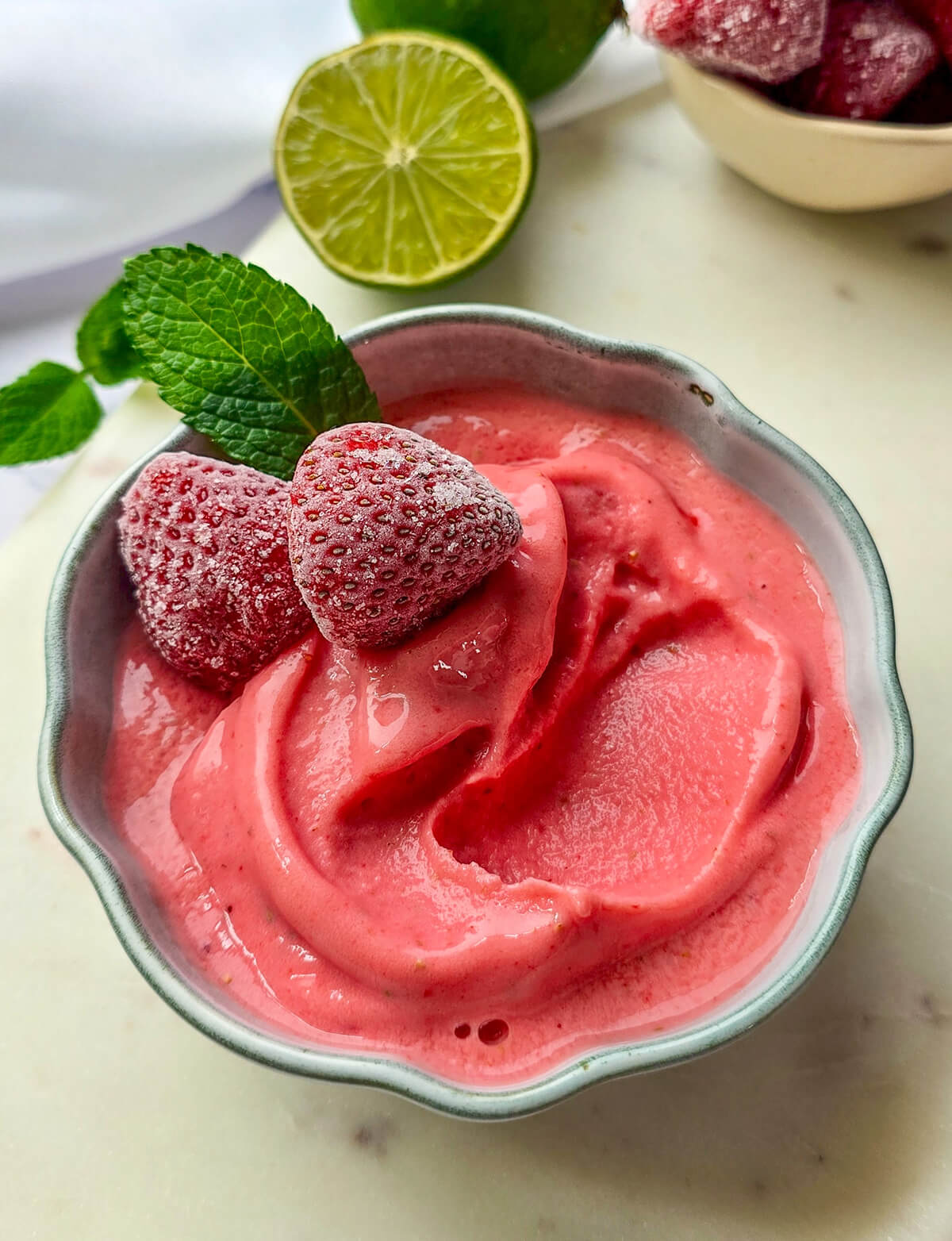Recette de sorbet fraise maison facile et rapide à faire sans sorbetière. Vue de dessus d'un bol de glace à la fraise onctueuse, décoré de fraises givrées et de menthe fraîche, avec des citrons verts en arrière-plan.