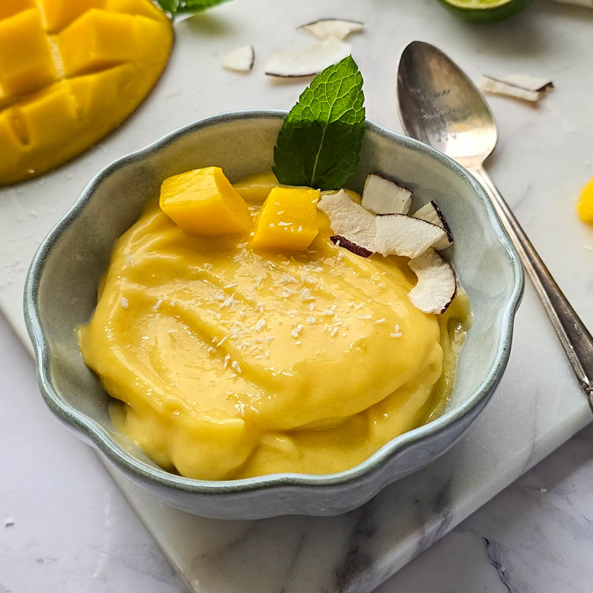Sorbet mangue maison rapide et rafraîchissant, réalisé sans sorbétière. Présentation du bol sur une planche en marbre avec une cuillère gravée Cuisine & Création et une mangue fraîche coupée à côté.