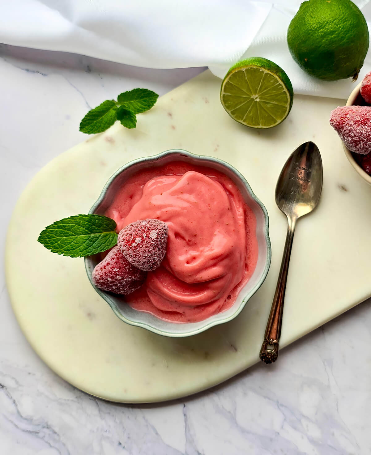 Sorbet fraise maison sans sorbetière, une idée de dessert glacé rapide et facile. Présentation sur une planche en marbre avec une cuillère gravée Cuisine & Création, de la menthe et des citrons verts.