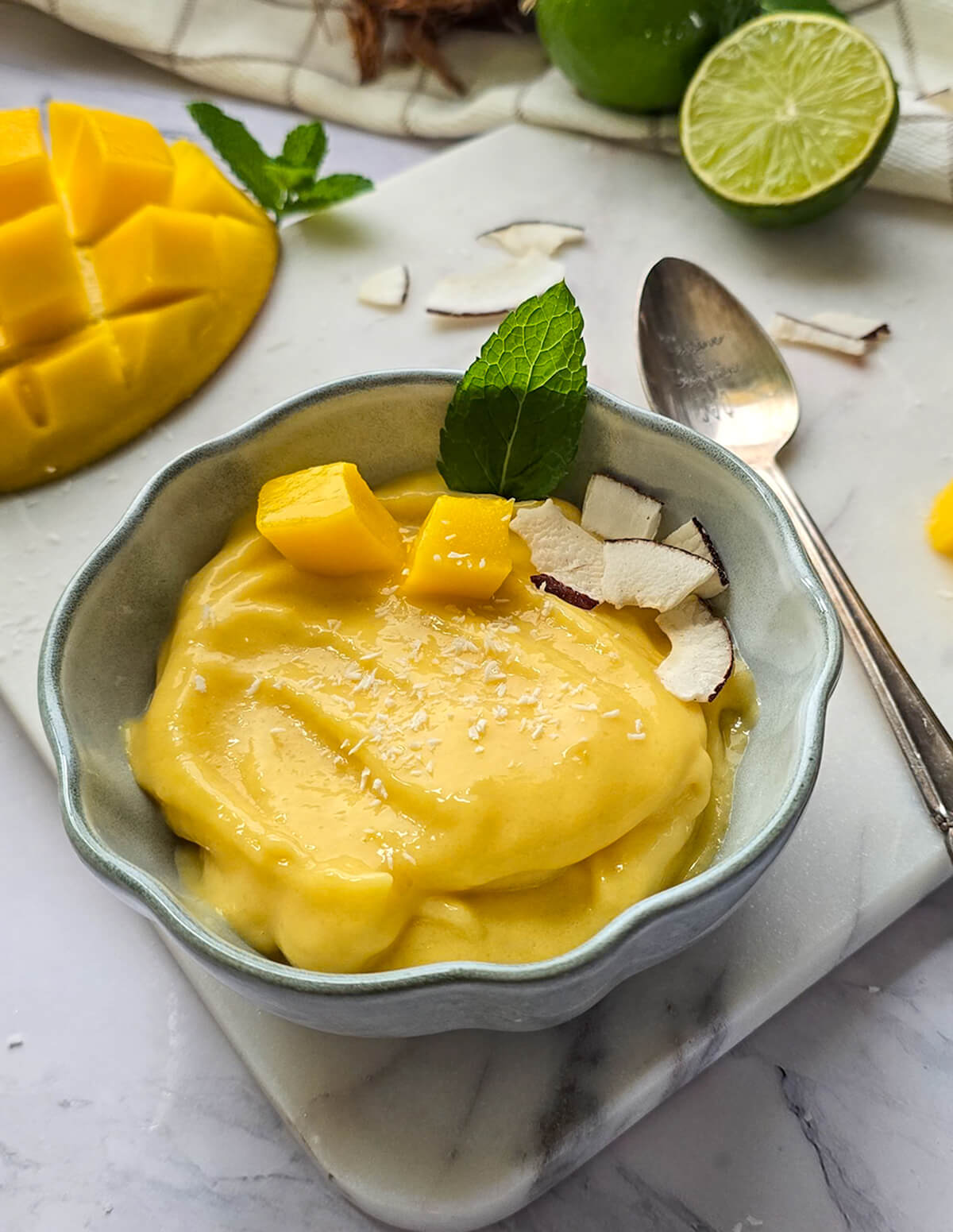 Recette facile de sorbet mangue maison sans sorbétière. Vue plongeante d'un bol de glace à la mangue décoré de dés de mangue fraîche, de copeaux de noix de coco et d'une feuille de menthe.