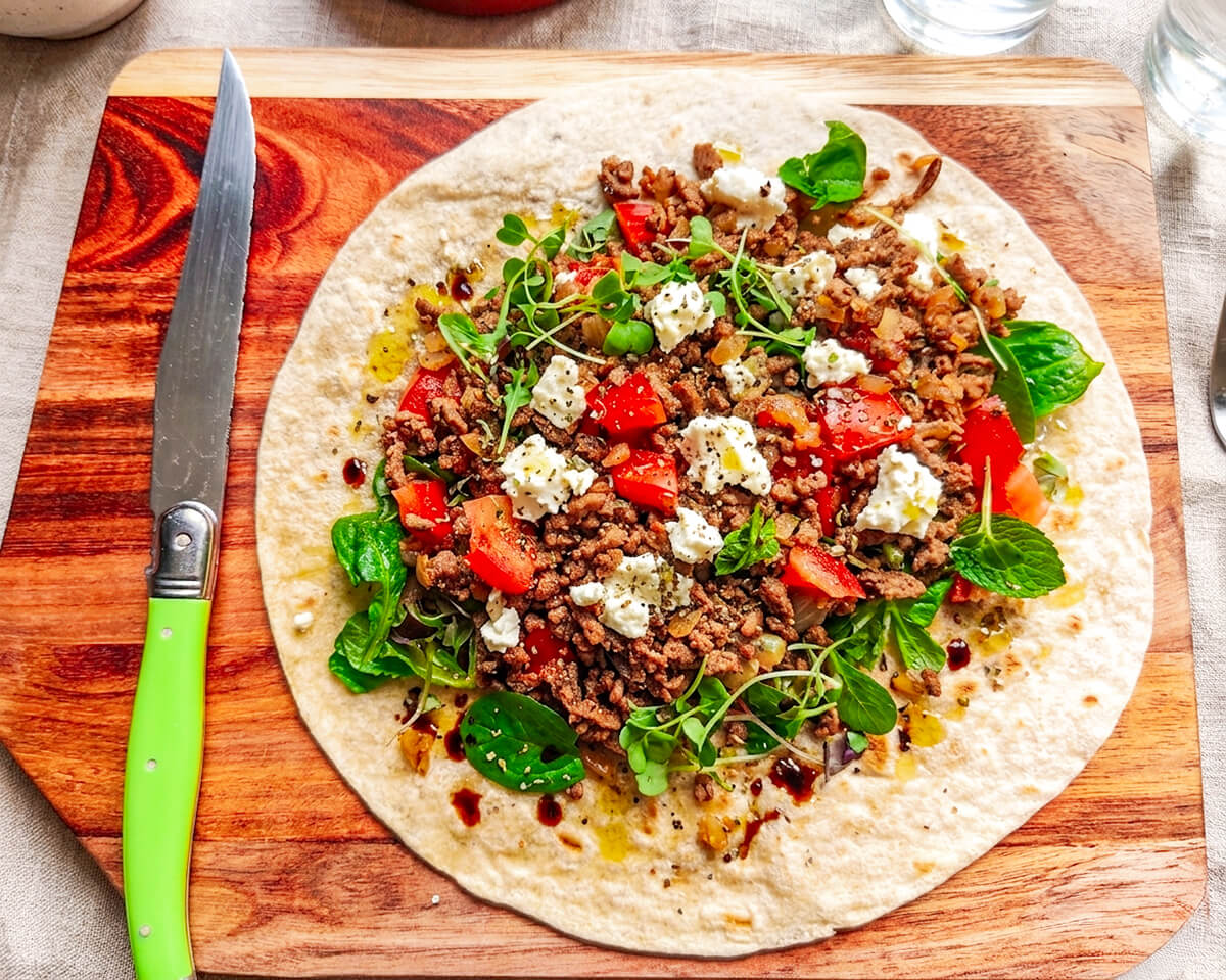 Un tacos de boeuf haché avec de la feta, tomates, mâche, couteau vert sur le côté, sur une planche de bois.