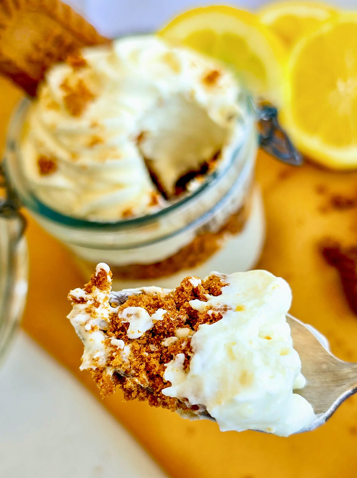Gros plan sur une cuillère en métal contenant une bouchée de verrine composée de crème onctueuse et de brisures de biscuits speculoos. En arrière-plan flou, on devine la verrine entamée et des tranches de citron jaune.