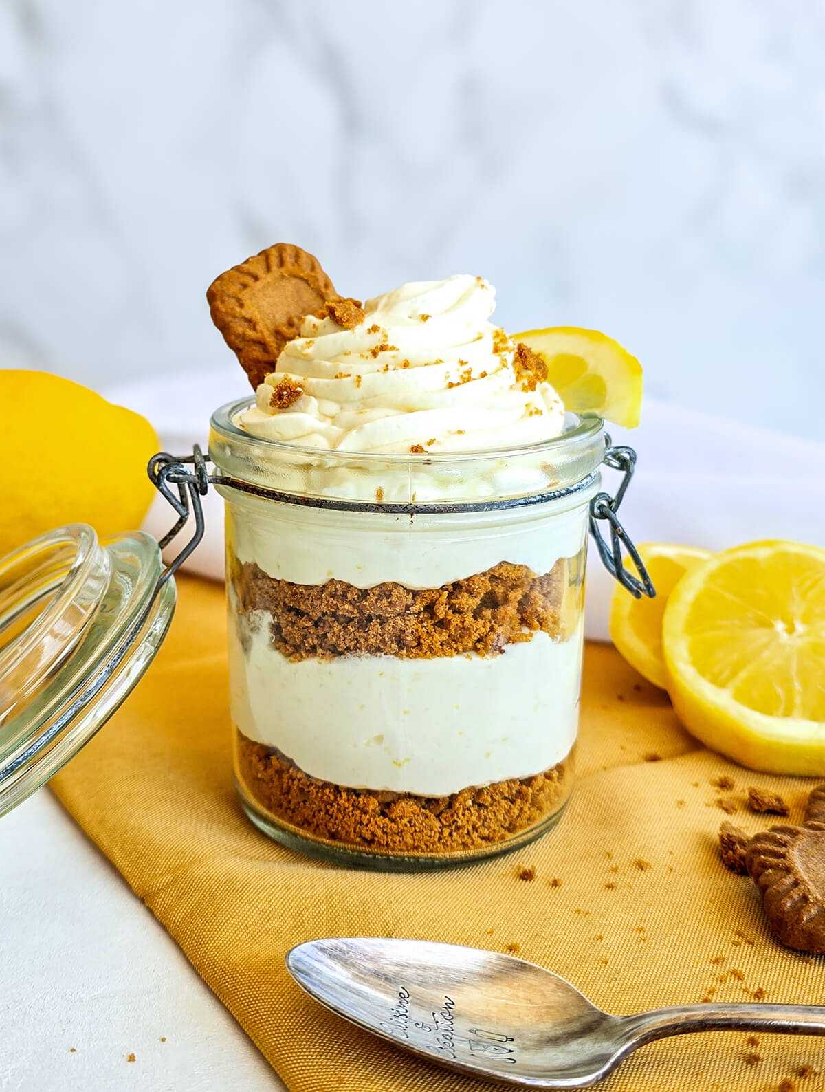 Vue d'ensemble de la verrine citron-speculoos posée sur un tissu jaune. On y voit clairement l'alternance des couches de biscuit brun et de crème blanche. Au premier plan, une cuillère en argent gravée est posée sur la table, et des citrons frais ainsi que des biscuits décorent l'arrière-plan.