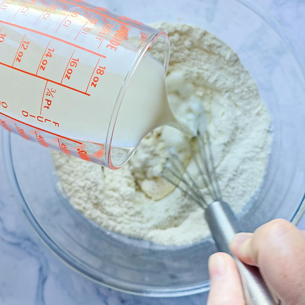 Une main mélangeant au fouet une pâte à pancakes, et une autre main versant du lait dans cette même pâte.