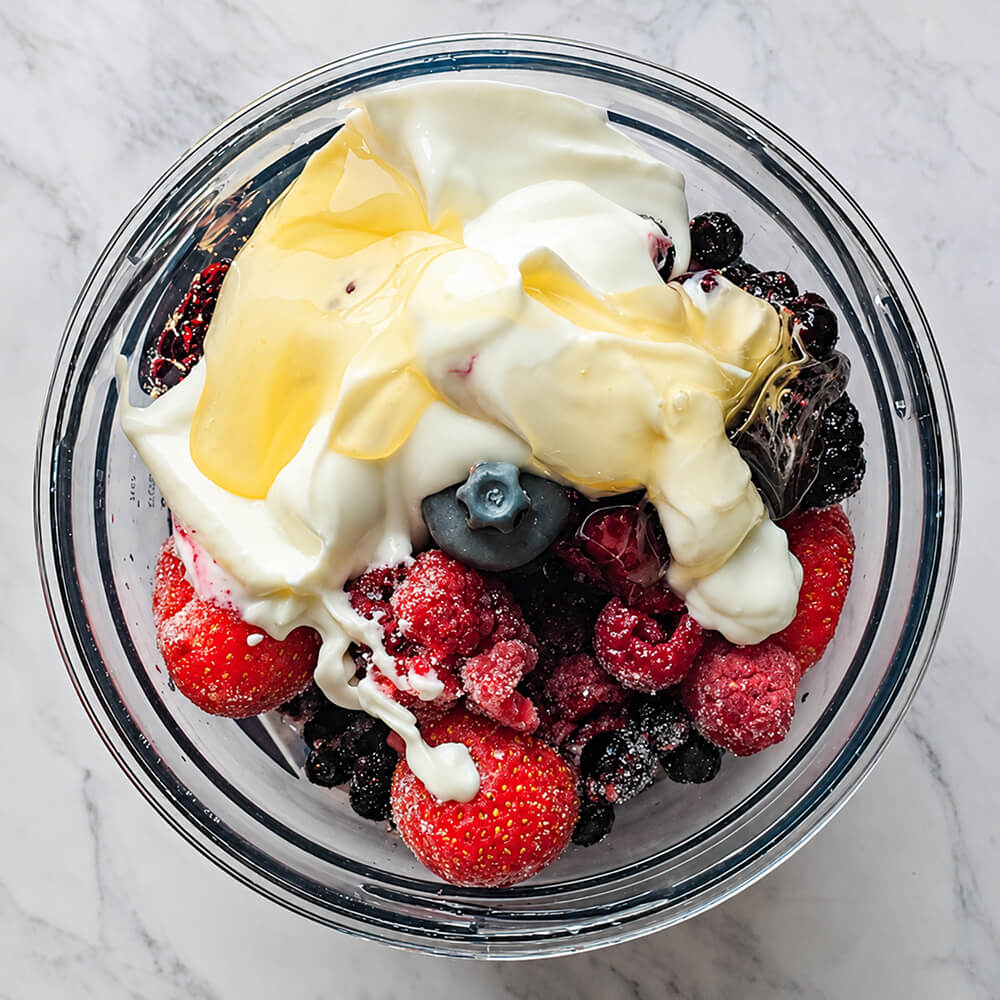 Des fruits rouges, du yaourt blanc et du miel dans le bol d'un petit mixeur, sur un fond blanc marbé pour la préparation d'une glace sans sorbetière.