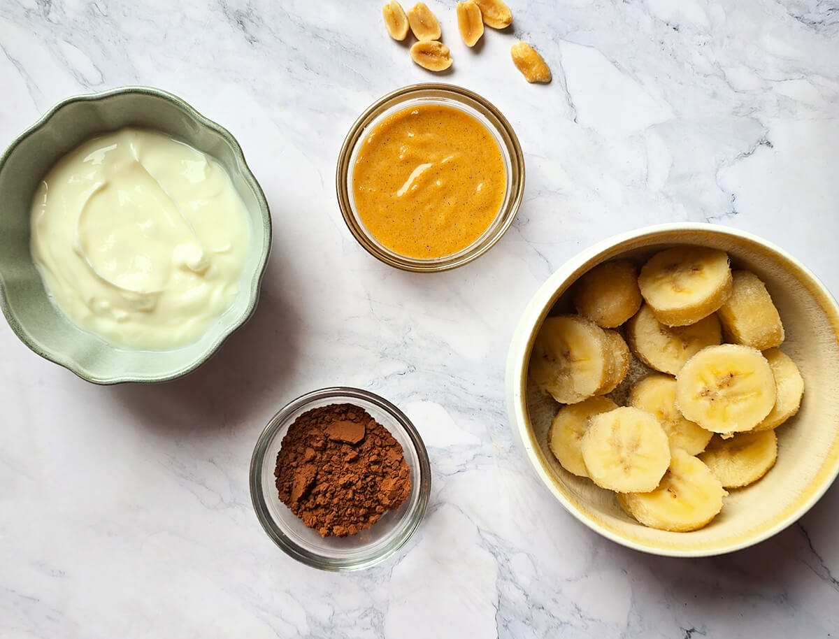 Ingrédients pour milkshake Snickers maison : yaourt grec, beurre de cacahuète, cacao en poudre et morceaux de banane congelée.