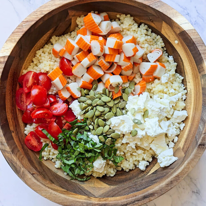 Grand saladier en bois contenant des perles de blé garnies de tomates cerises coupées, dés de surimi, feta, graines de courge et basilic ciselé.