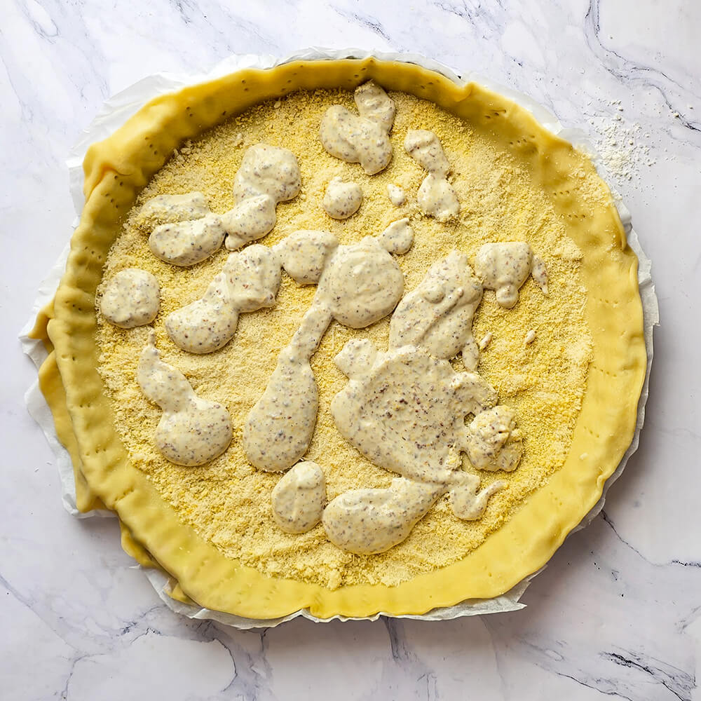 Une tarte non cuite dans son moule avec du papier sulfurisé. On peut voir de la chapelure et un mélange de moutarde et crème, sur fond blanc marbré.