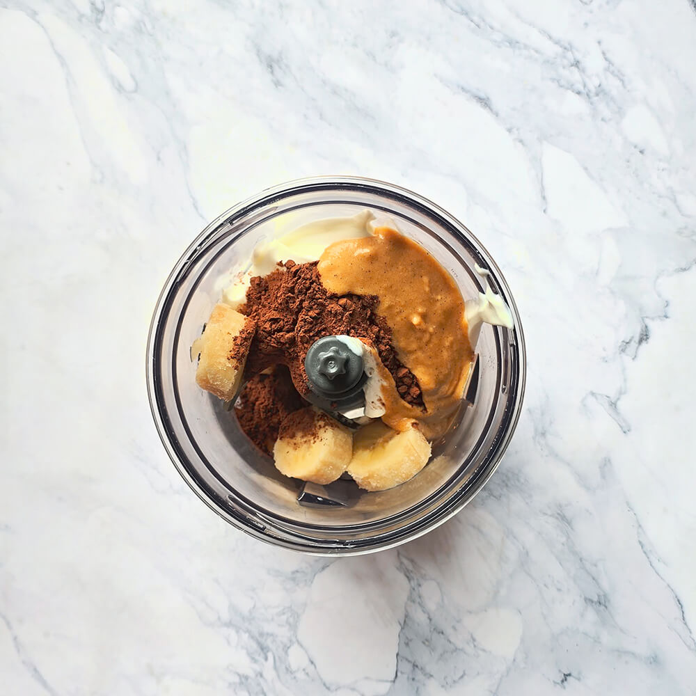 Préparation d'un milkshake Snickers : yaourt, beurre de cacahuète, cacao et banane dans un mixeur sur un plan de travail en marbre.