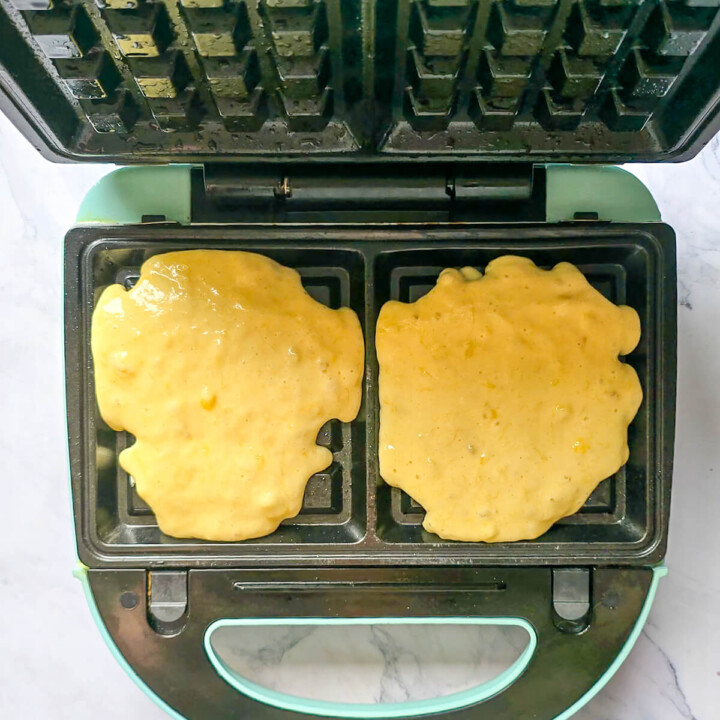 Louches de pâte à gaufre à la banane déposées dans les empreintes d'un gaufrier électrique ouvert d'une couleur bleu pastel.
