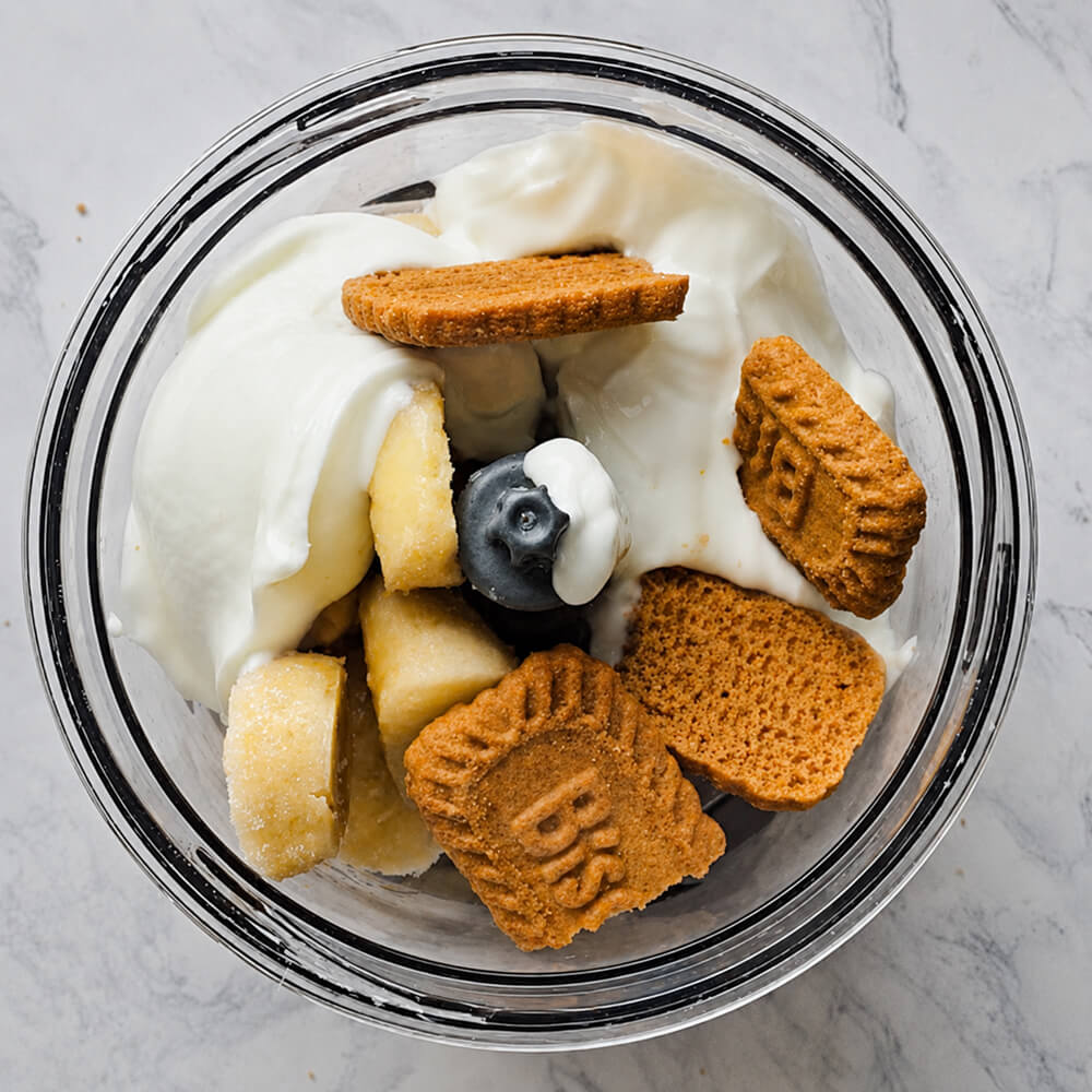 Vue plongeante dans le bol d'un mixeur contenant des rondelles de bananes surgelées, des morceaux de biscuits Biscoff et une généreuse portion de fromage blanc, prêts à être mixés.