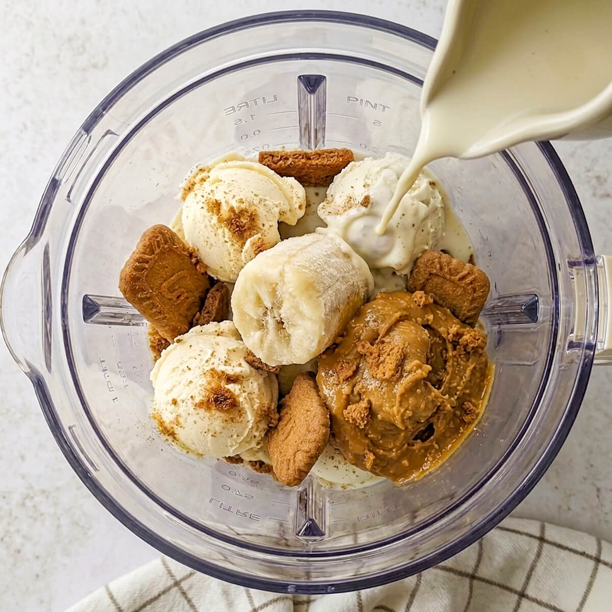 Vue plongeante sur un mixeur contenant des morceaux de banane, des boules de glace, de la pâte à tartiner spéculoos et un biscuit spéculoos coupé en deux, tandis qu'une main y verse du lait.