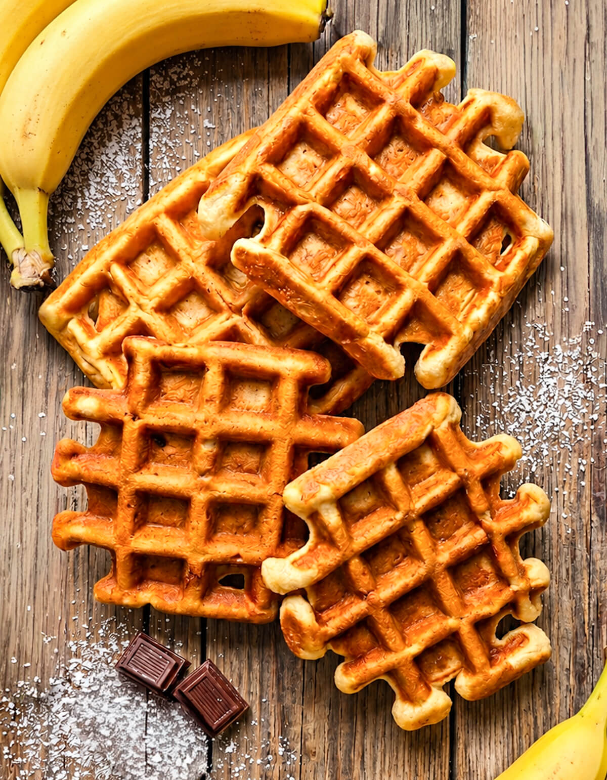 Vue de dessus de quatre gaufres banane dorées sur une table en bois avec une banane et du chocolat.