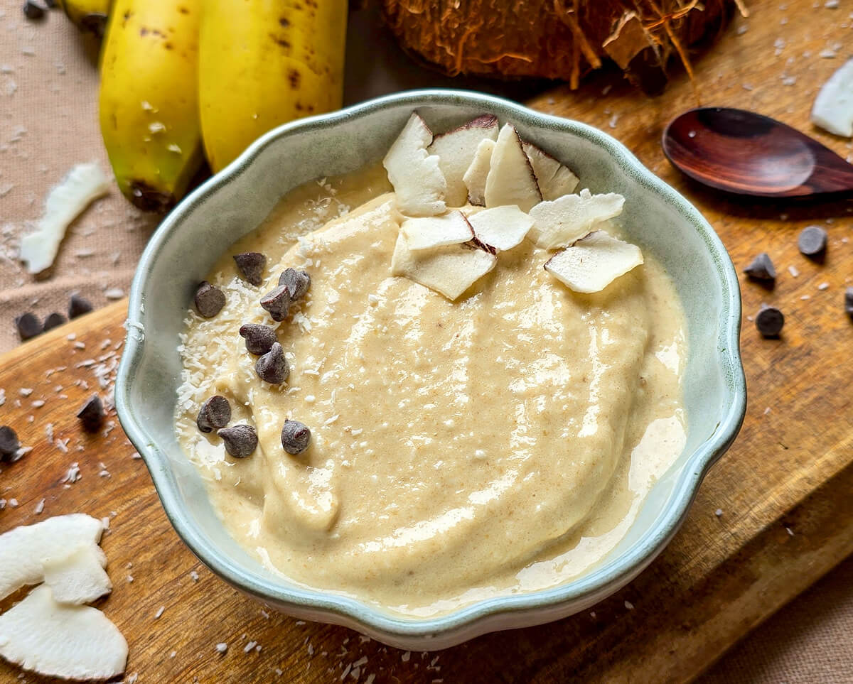 Bol de glace onctueuse banane-coco décoré de copeaux de noix de coco fraîche et de pépites de chocolat noir, posé sur une planche en bois rustique avec deux bananes mûres et une noix de coco entière en arrière-plan.