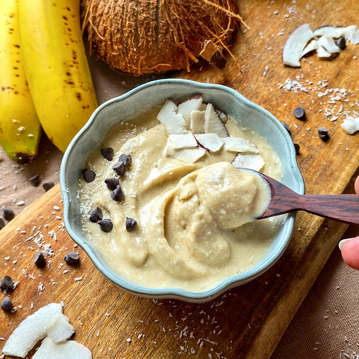 Cuillère en bois avec de la glace coco-banane dans un bol blanc sur une planche en bois, accompagnée de noix de coco râpée, de pépites de chocolat et de banane en arrière-plan.