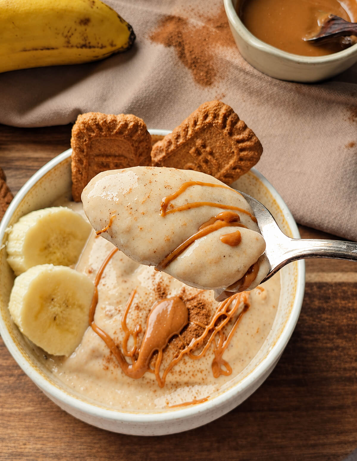 Une cuillère soulève une bouchée onctueuse de glace à la banane, sur laquelle coule un filet de pâte à tartiner spéculoos. La cuillère est positionnée au-dessus d'un bol contenant le reste de la glace, garni de rondelles de banane fraîche et de biscuits Lotus Biscoff entiers. L'arrière-plan comprend une banane entière et d'autres biscuits sur une surface en bois.