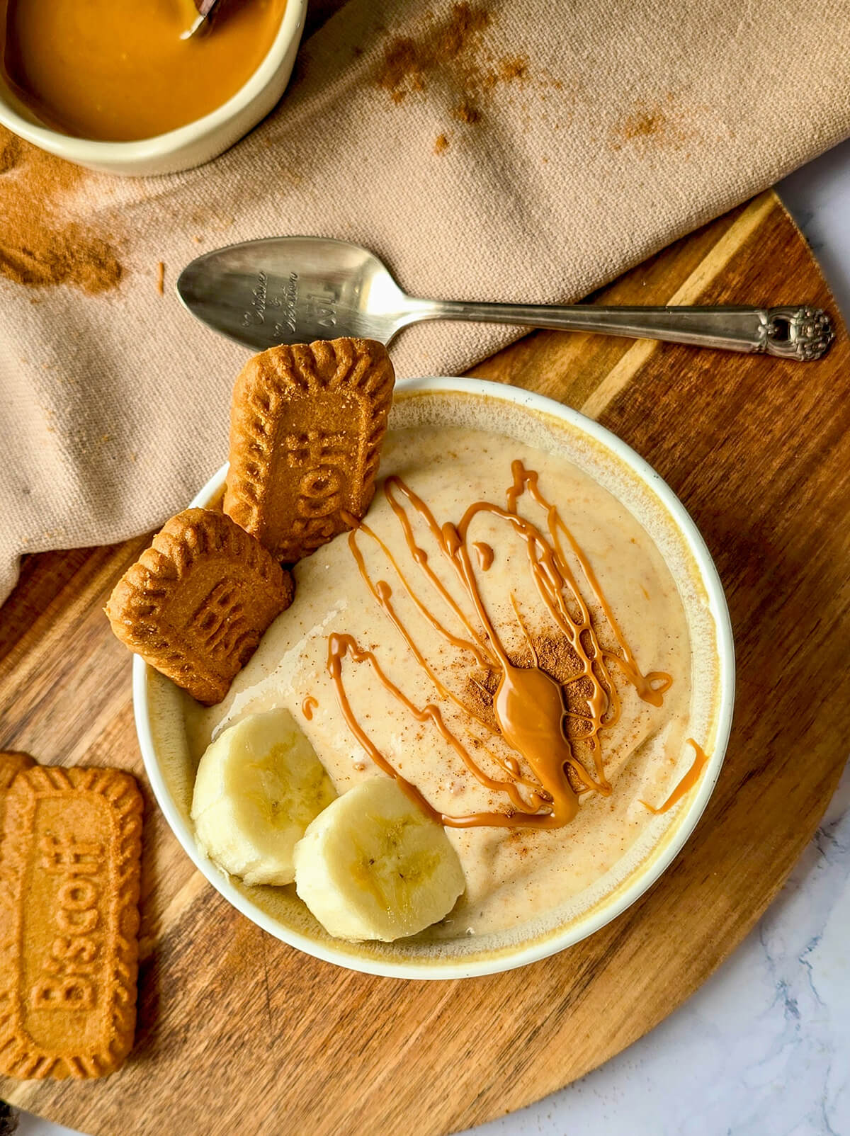 Un bol de glace à la banane, posé sur une planche à découper en bois et garni de tranches de banane, de deux biscuits Lotus Biscoff et d'un filet de sauce au caramel, ainsi que d'une pincée de cannelle.