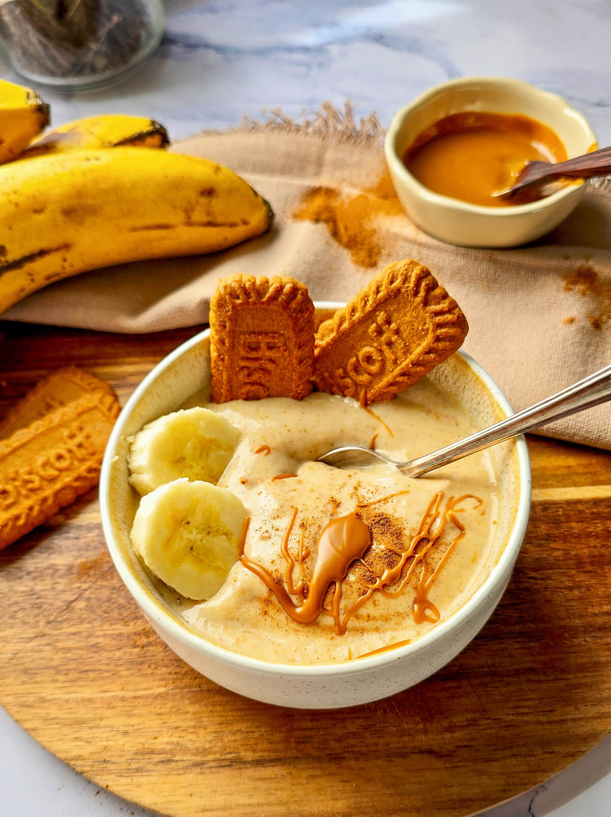Bol de glace à la banane onctueuse, garni de deux biscuits Biscoff, de rondelles de banane fraîche, d'un filet de beurre de spéculoos et d'une touche de cannelle, présenté sur une planche en bois avec des bananes entières en arrière-plan.