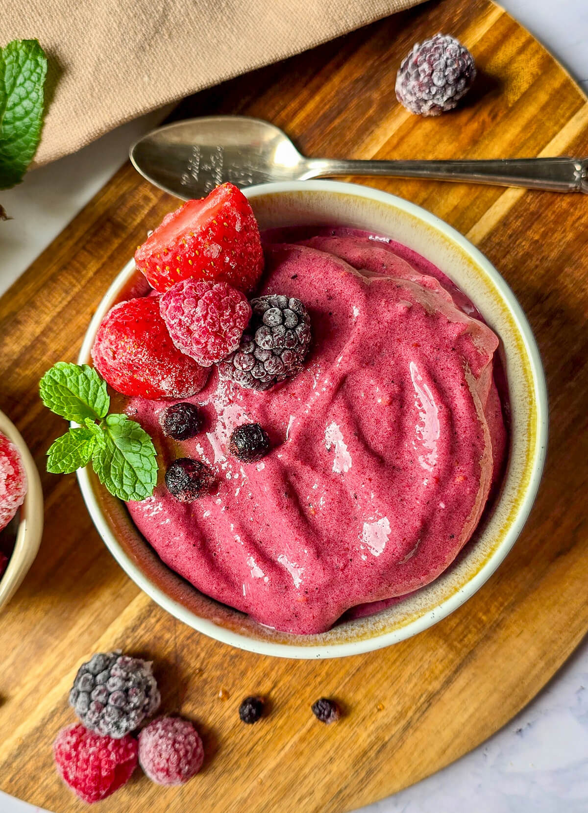 Gros plan sur une glace de couleur rose foncé, avec des fruits rouges surgelés, sur une planche de bois.