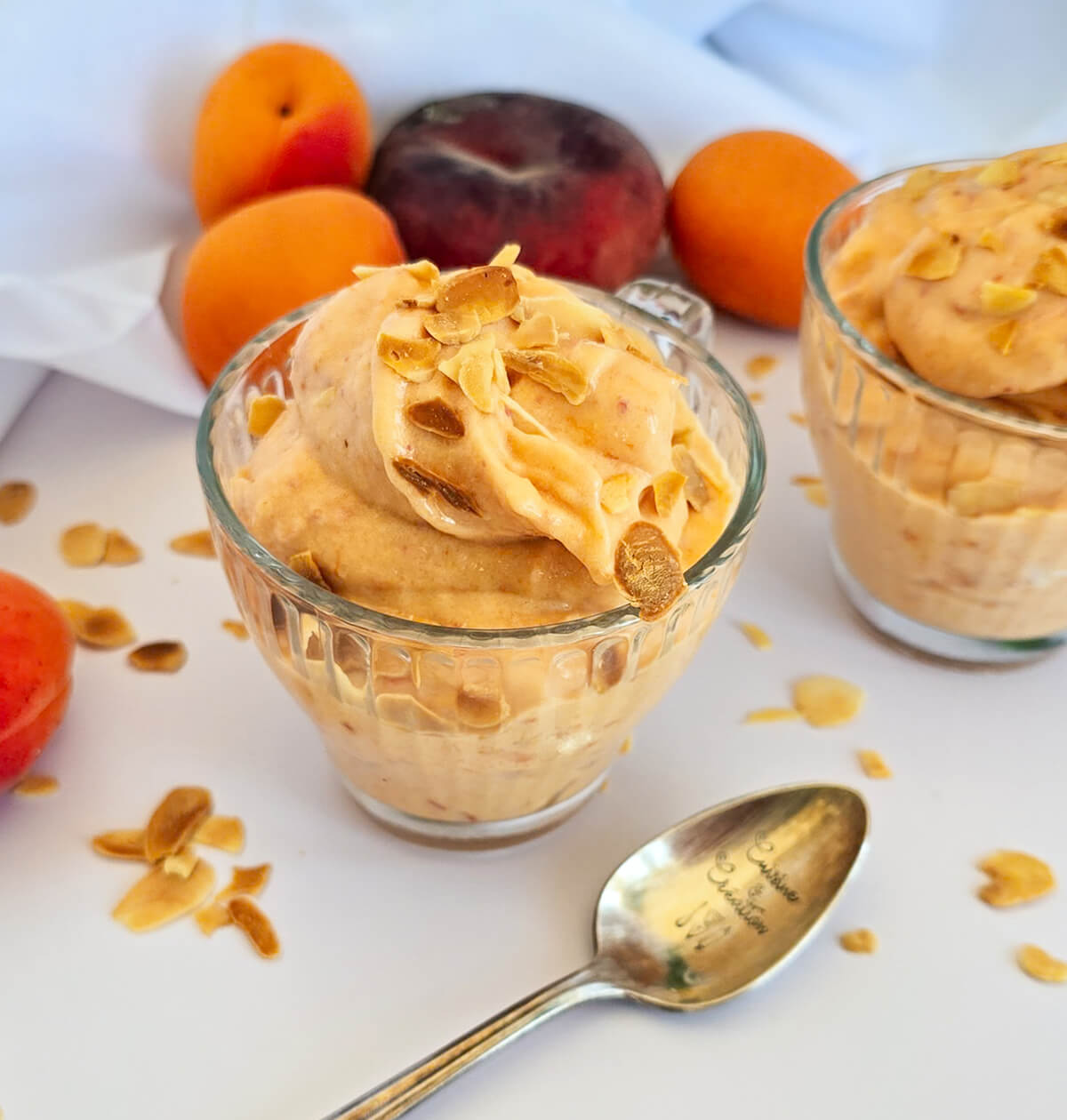 Deux coupes en verre remplies de glace maison à la pêche et à l'abricot sans sorbetière, décorées d'amandes effilées, avec une cuillère gravée Cuisine et Création au premier plan.