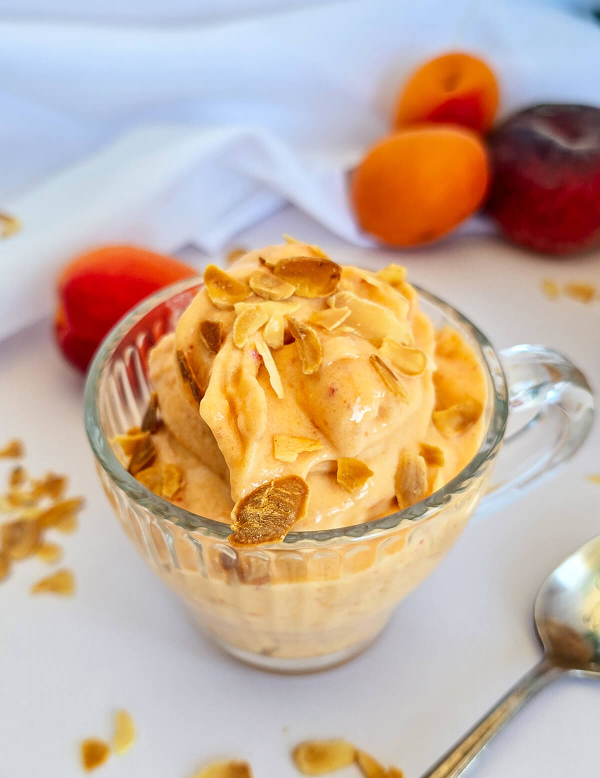 Une coupe en verre givrée remplie d'une glace onctueuse à la pêche et à l'abricot, parsemée d'amandes effilées grillées, avec des fruits frais en arrière-plan.