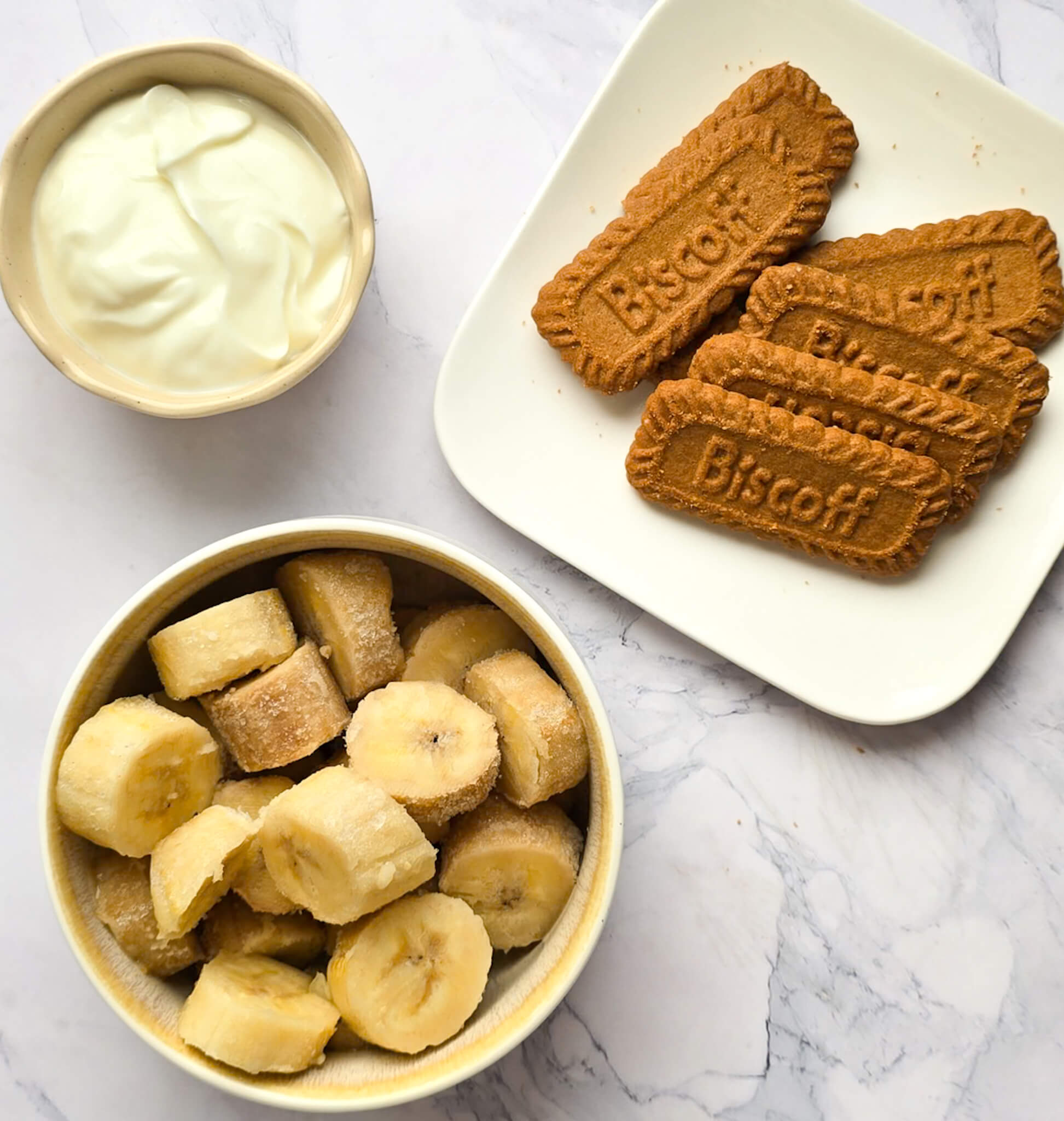 Vue de dessus des ingrédients pour une glace à la banane et au spéculoos : un bol de rondelles de bananes surgelées, une assiette avec plusieurs biscuits Biscoff et un petit bol de crème onctueuse sur un fond marbré blanc.