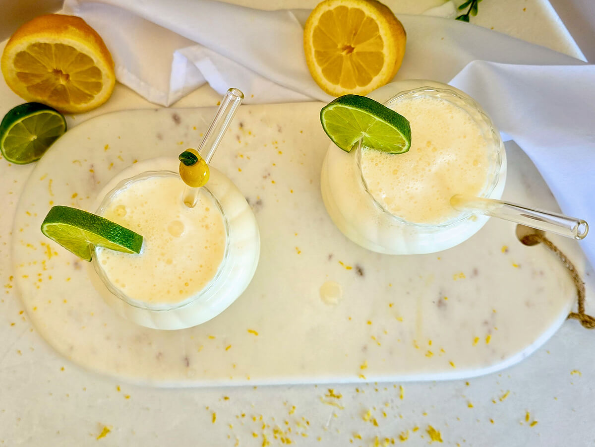 Deux verres de limonade crémeuse brésilienne vus du dessus, posés sur une planche en marbre blanc parsemée de zestes, décorés de rondelles de citron vert et de pailles en verre.