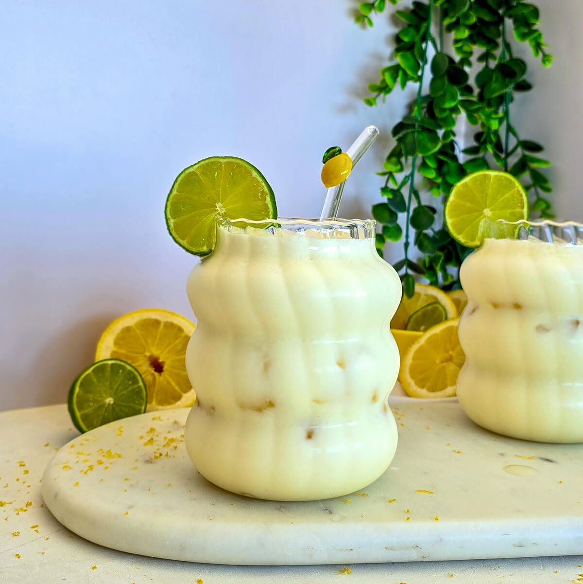 Vue de face d'un verre ondulé transparent rempli de limonade crémeuse au citron vert avec des glaçons, surélevé sur un plateau en marbre avec des citrons en arrière-plan.