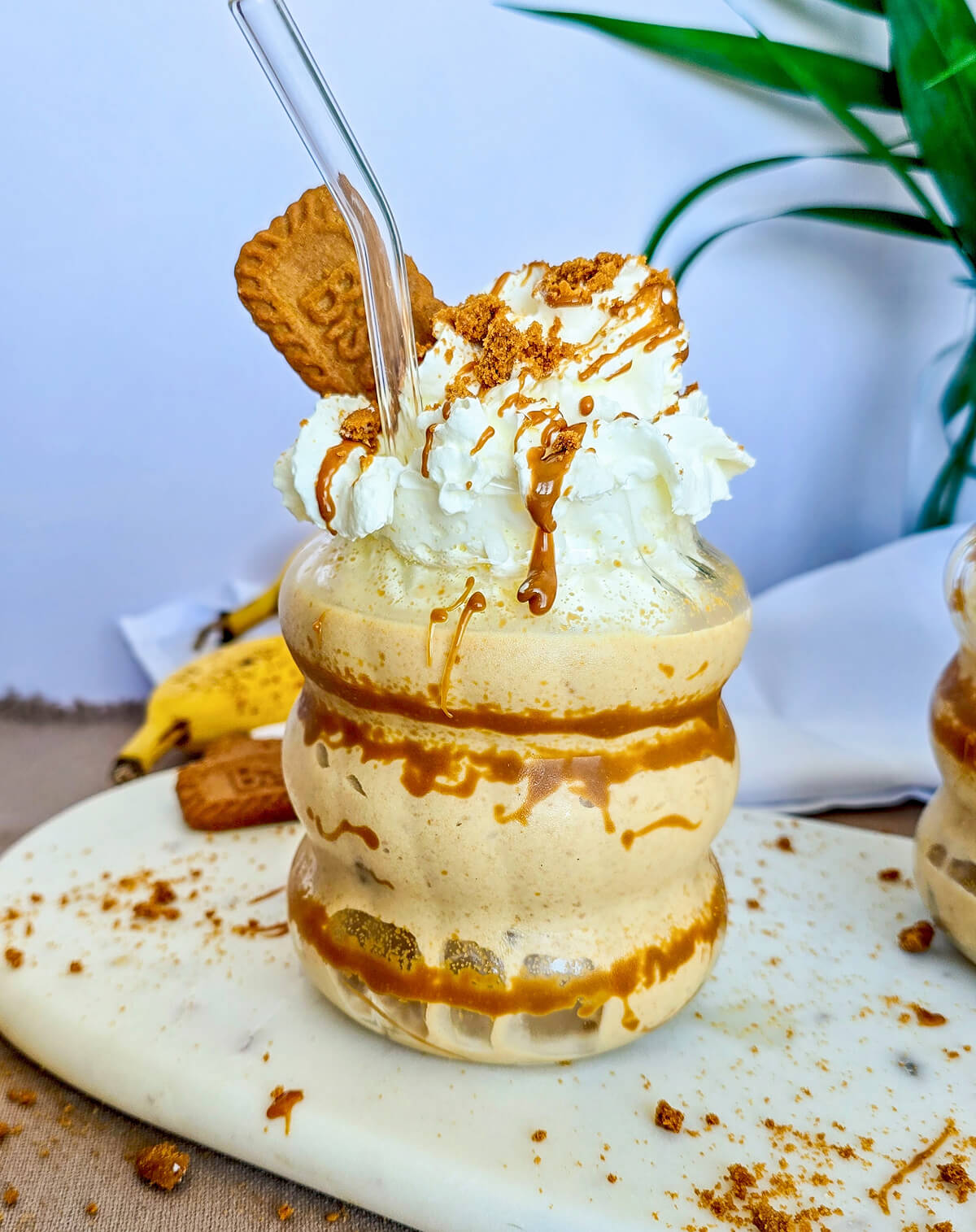 Milkshake banane et spéculoos dans un verre ondulé, nappé de coulant de spéculoos, surmonté de crème chantilly, d'un biscuit Biscoff et d'une paille en verre.