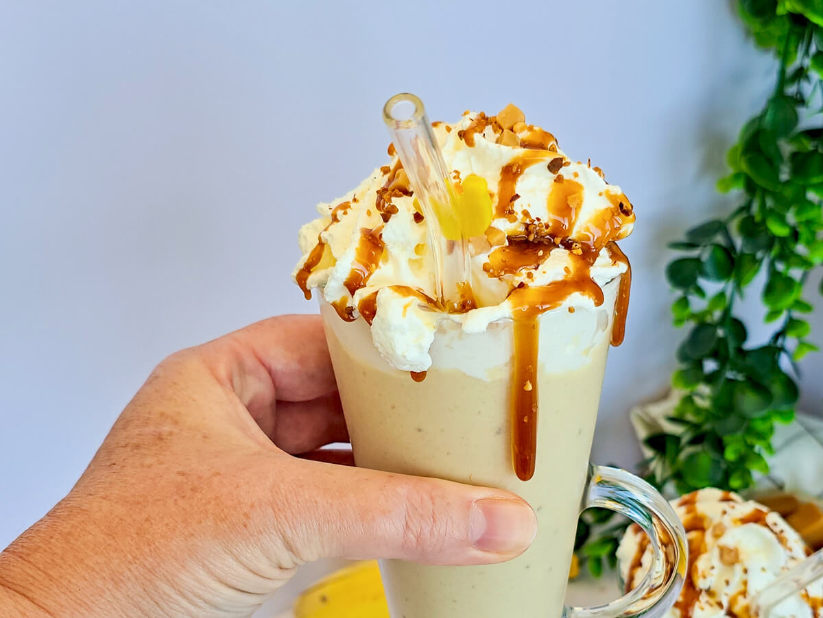 Un milkshake banane vanille dans un grand verre, surmonté de crème chantilly, d'un coulage de caramel et d'éclats de spéculoos, tenu par une main.