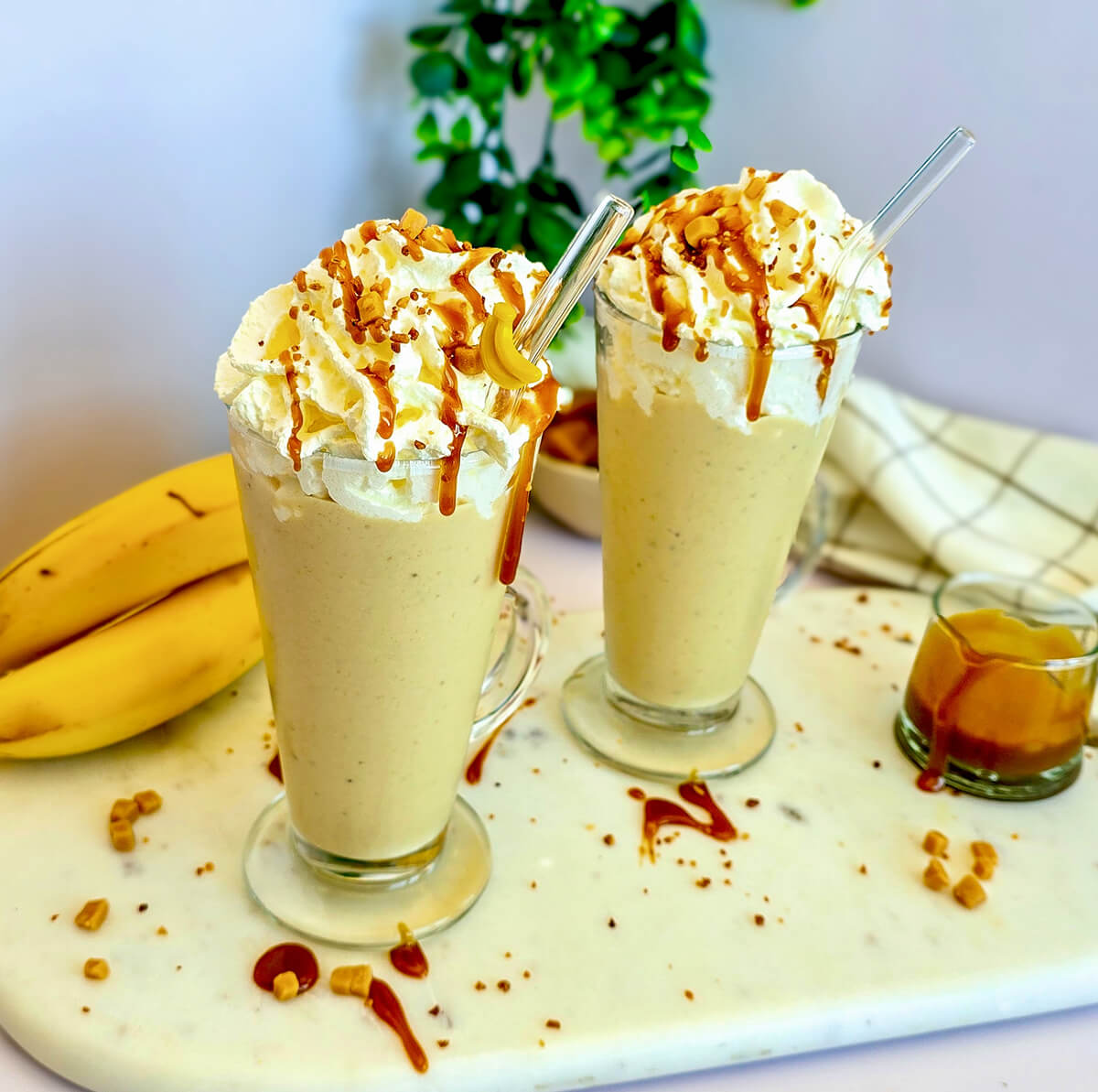 Deux verres de milkshake crémeux au caramel et à la banane sur un plateau en marbre, décorés de crème fouettée, de sauce caramel et de morceaux de banane. À l'arrière-plan, des bananes entières, une petite tasse de sauce caramel et une serviette de table.