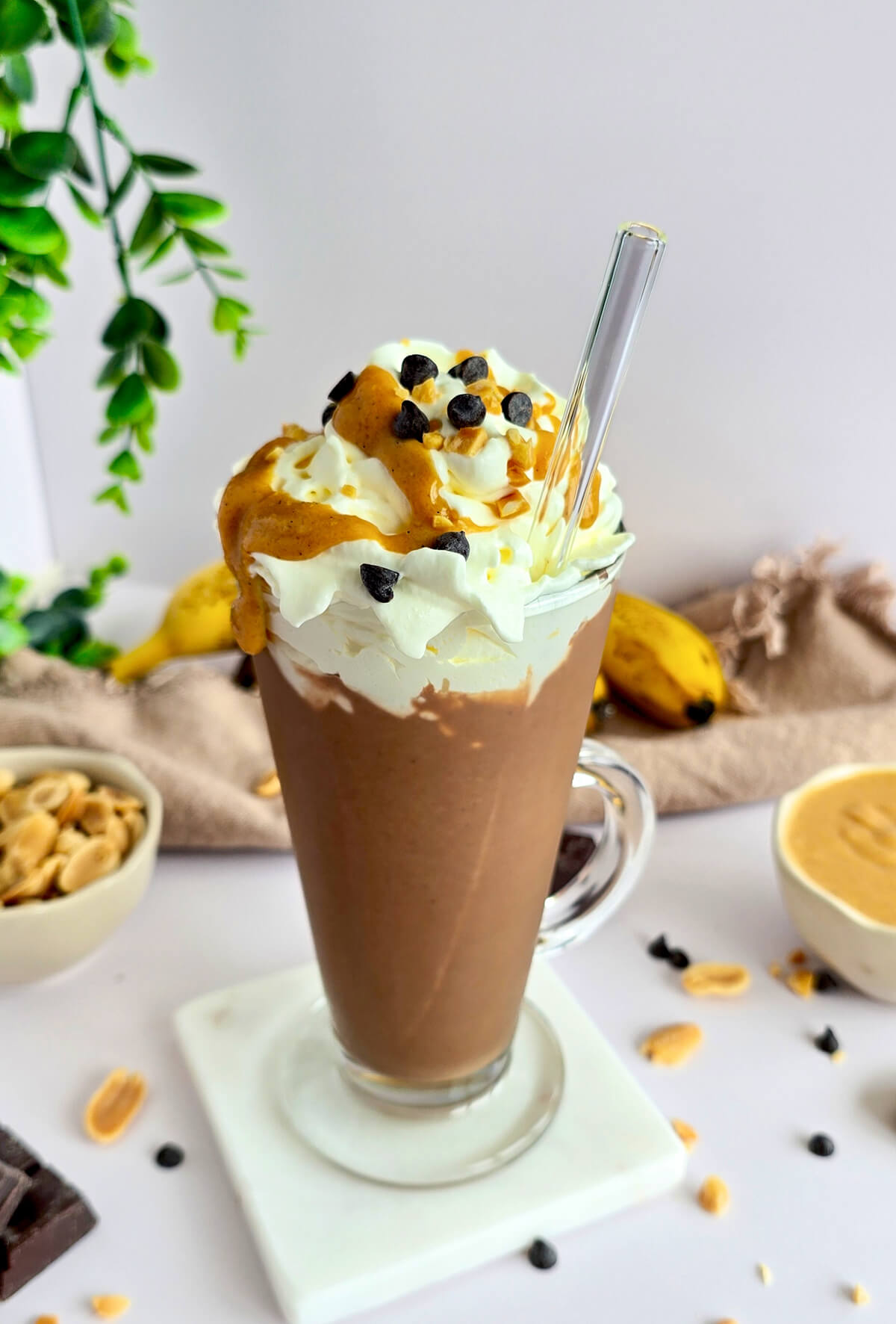 Milkshake Snickers facile dans un grand verre avec paille, garni de chantilly, caramel et pépites de chocolat, entouré d'ingrédients.