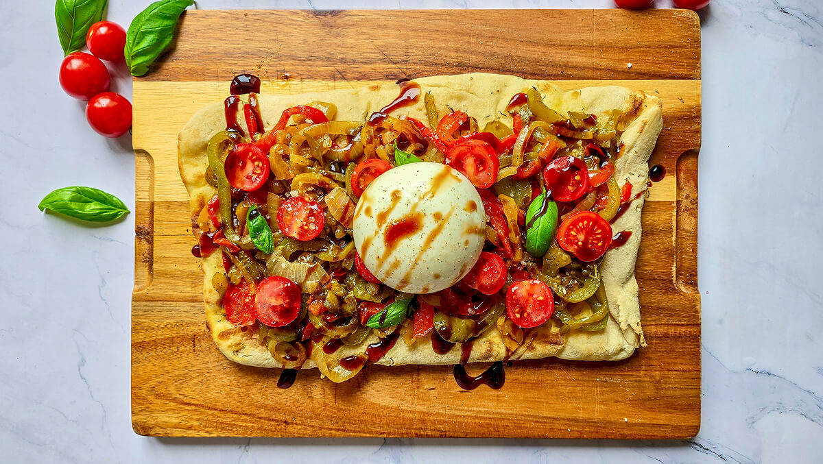 Pinsa garnie de poivrons sautés, tomates cerises et burrata entière, agrémentée d'un trait de crème de vinaigre balsamique sur une planche en bois.