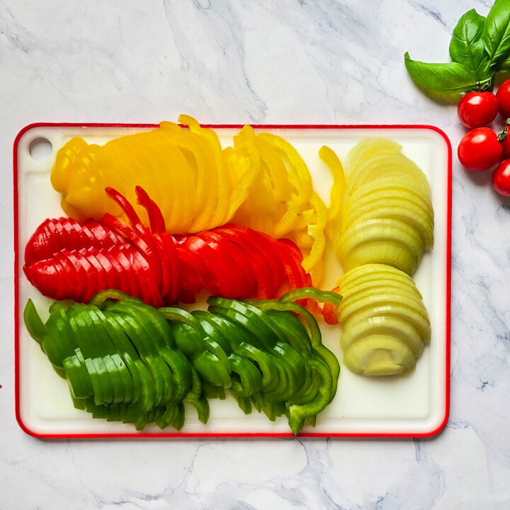 Planche à découper présentant des tranches fines de poivrons jaunes, rouges et verts, ainsi que des lamelles d'oignons prêtes à être cuisinées.