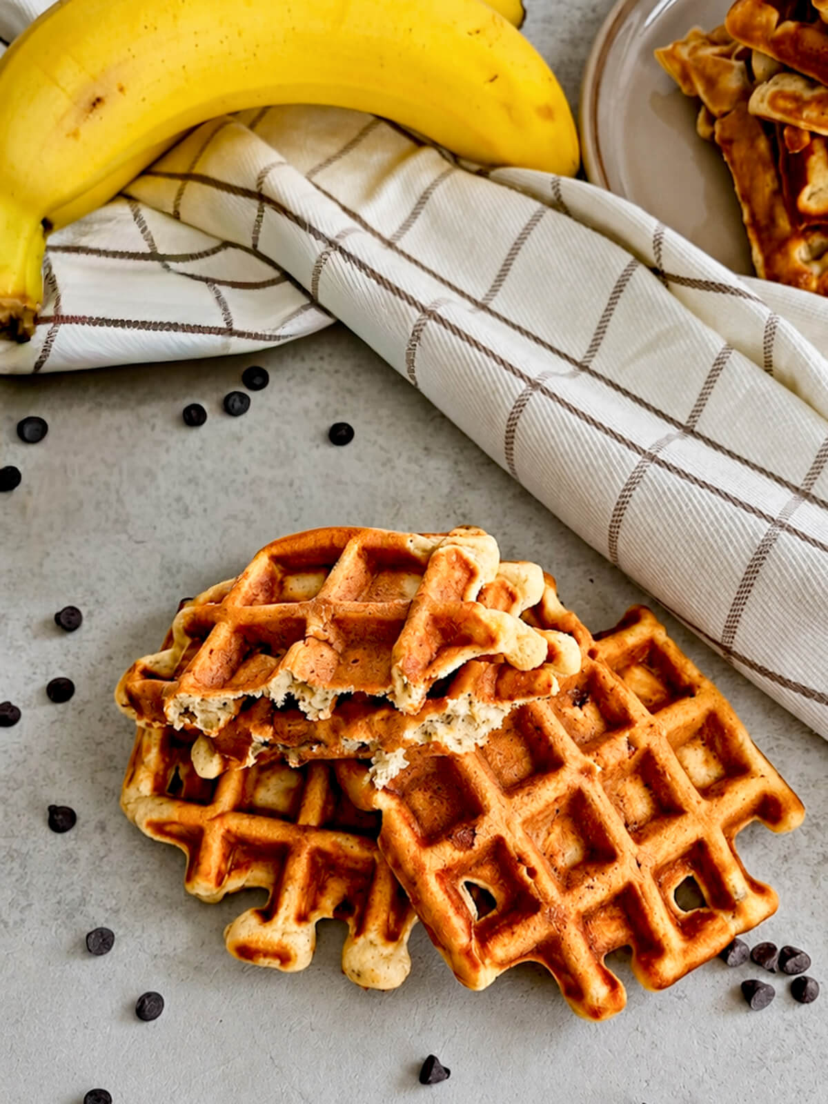 Gaufres à la banane faites maison et pépites de chocolat noir parsemées sur un plan de travail gris clair, avec des bananes fraîches et un torchon blanc à carreaux en arrière-plan.