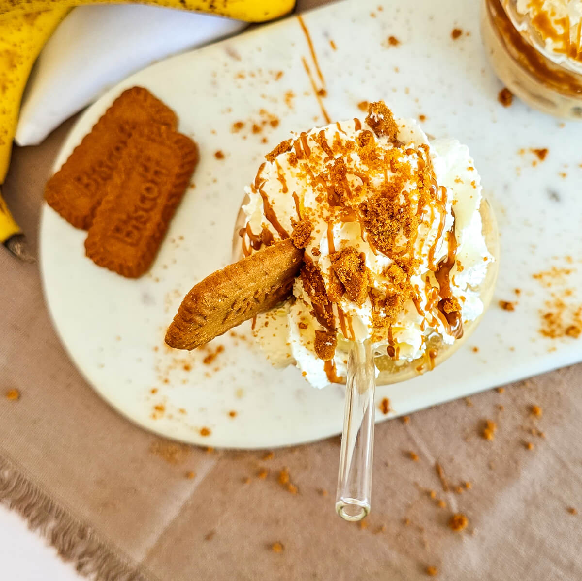 Vue du dessus d'un gourmand milkshake à la banane et au spéculoos avec chantilly, brisures de biscuits et coulis, posé sur une planche en marbre blanc.
