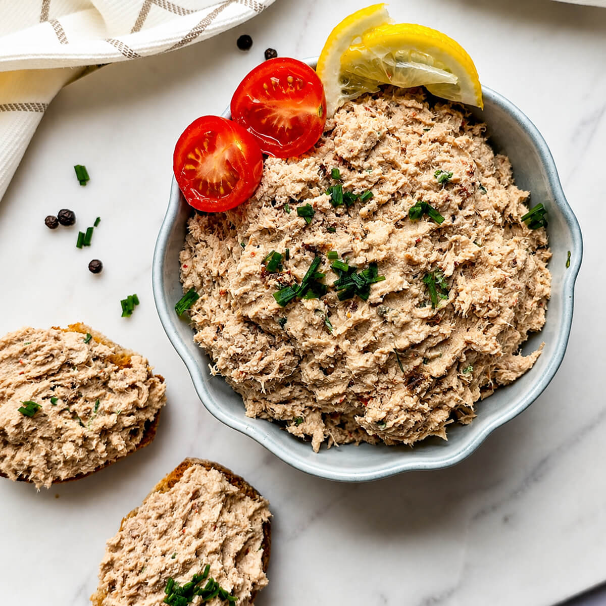 des rillettes de thon faciles et crémeuses dans un bol blanc avec une tomate cerise coupée en deux, et sur des tracnhes de pain, sur un fond blanc marbré,
