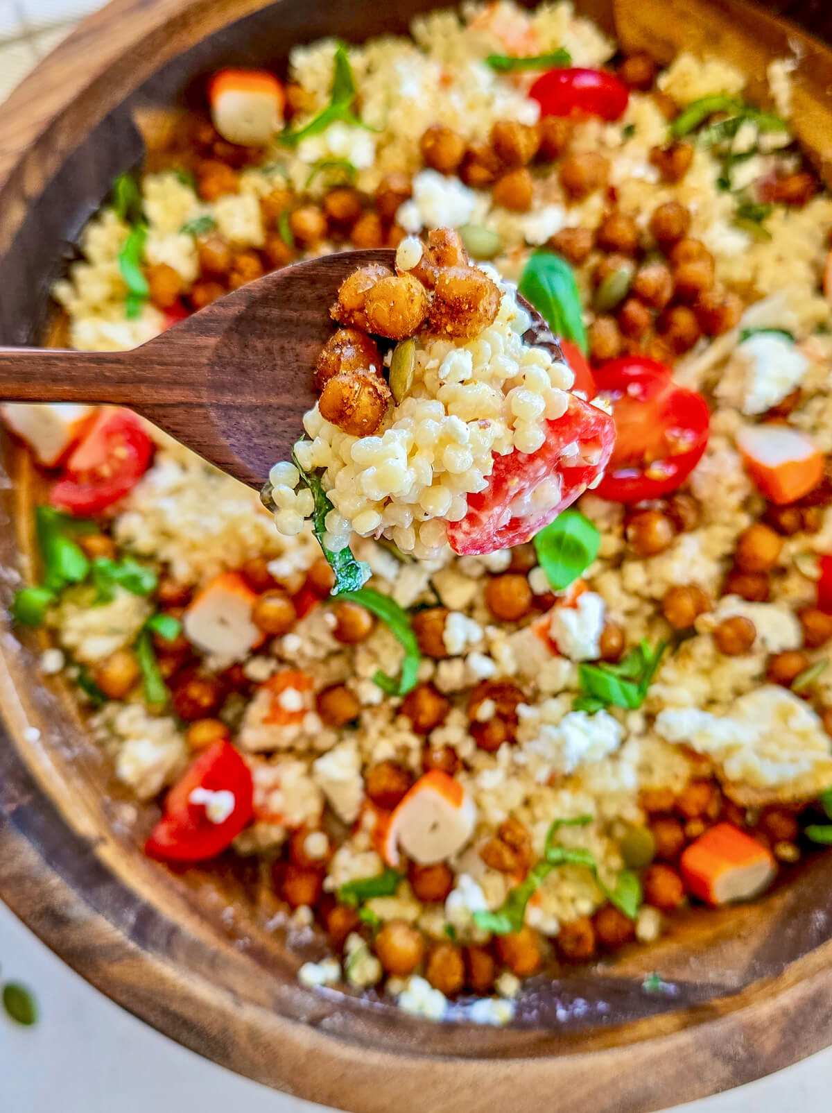 Gros plan en plongée d'une cuillère en bois prélevant une bouchée de salade de perles de blé, montrant la texture du couscous perlé, des pois chiches rôtis aux épices, de la feta émiettée, des tomates cerises et du basilic ciselé, avec le saladier en bois en arrière-plan flou.